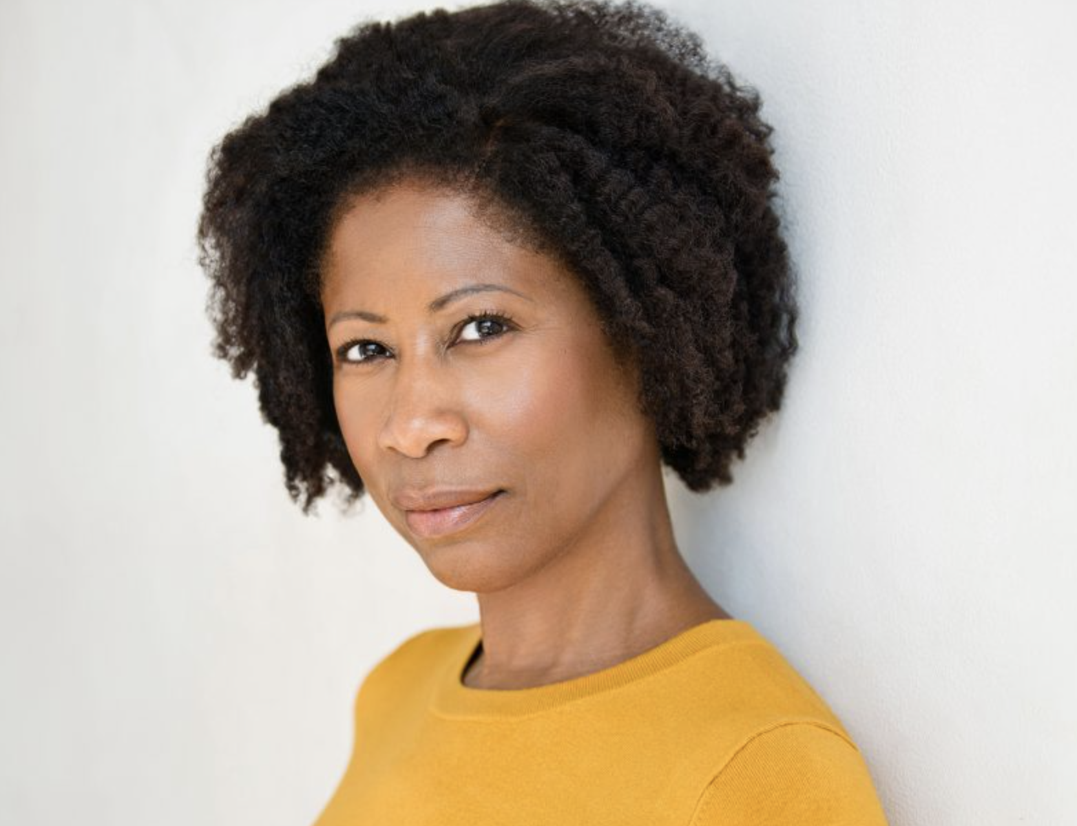 A woman with natural curly hair wearing a yellow top, looking slightly to the right, standing against a plain light-colored background.