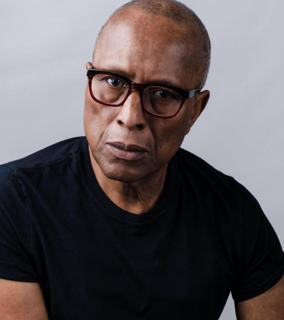 Close-up of a middle-aged man with a shaved head, wearing red-rimmed glasses and a black shirt, looking serious.
