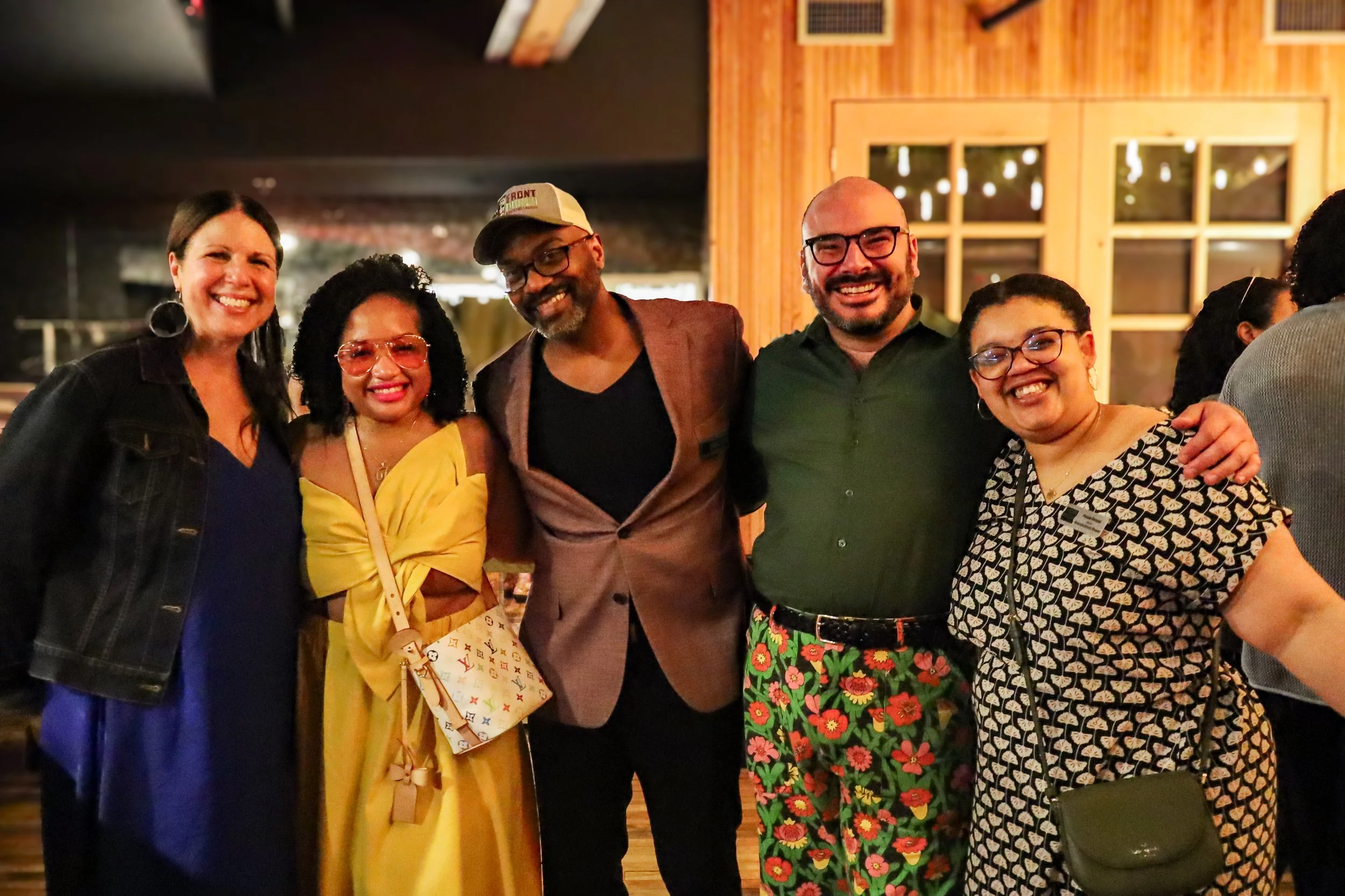 Five people standing together and smiling in a warmly lit indoor setting. The group includes women and men of diverse ethnicities, dressed in casual to semi-formal attire. The background features wooden walls and large windows with visible lights.