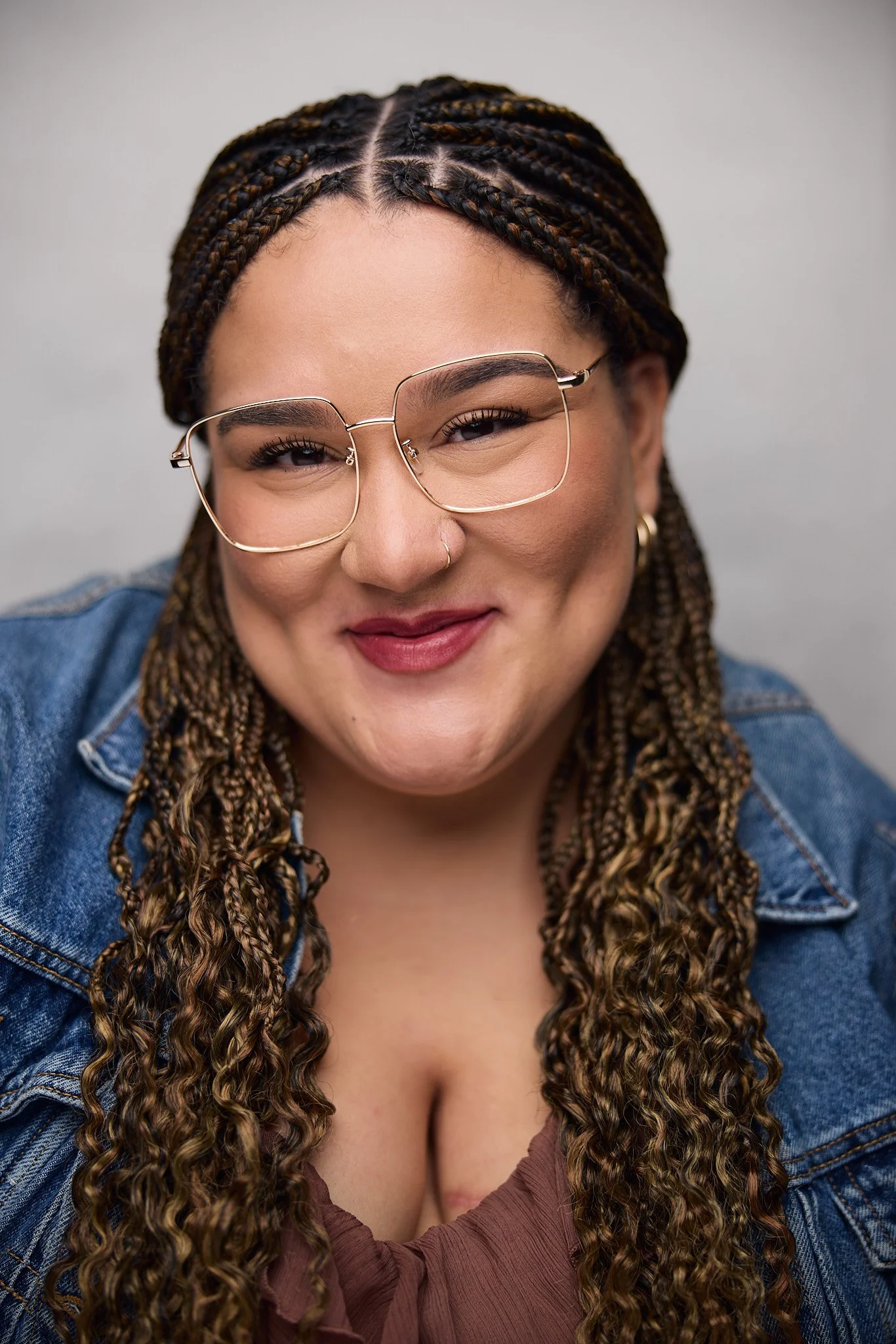 Close-up of a woman with braided hair wearing large, square glasses and a denim jacket, smiling softly.