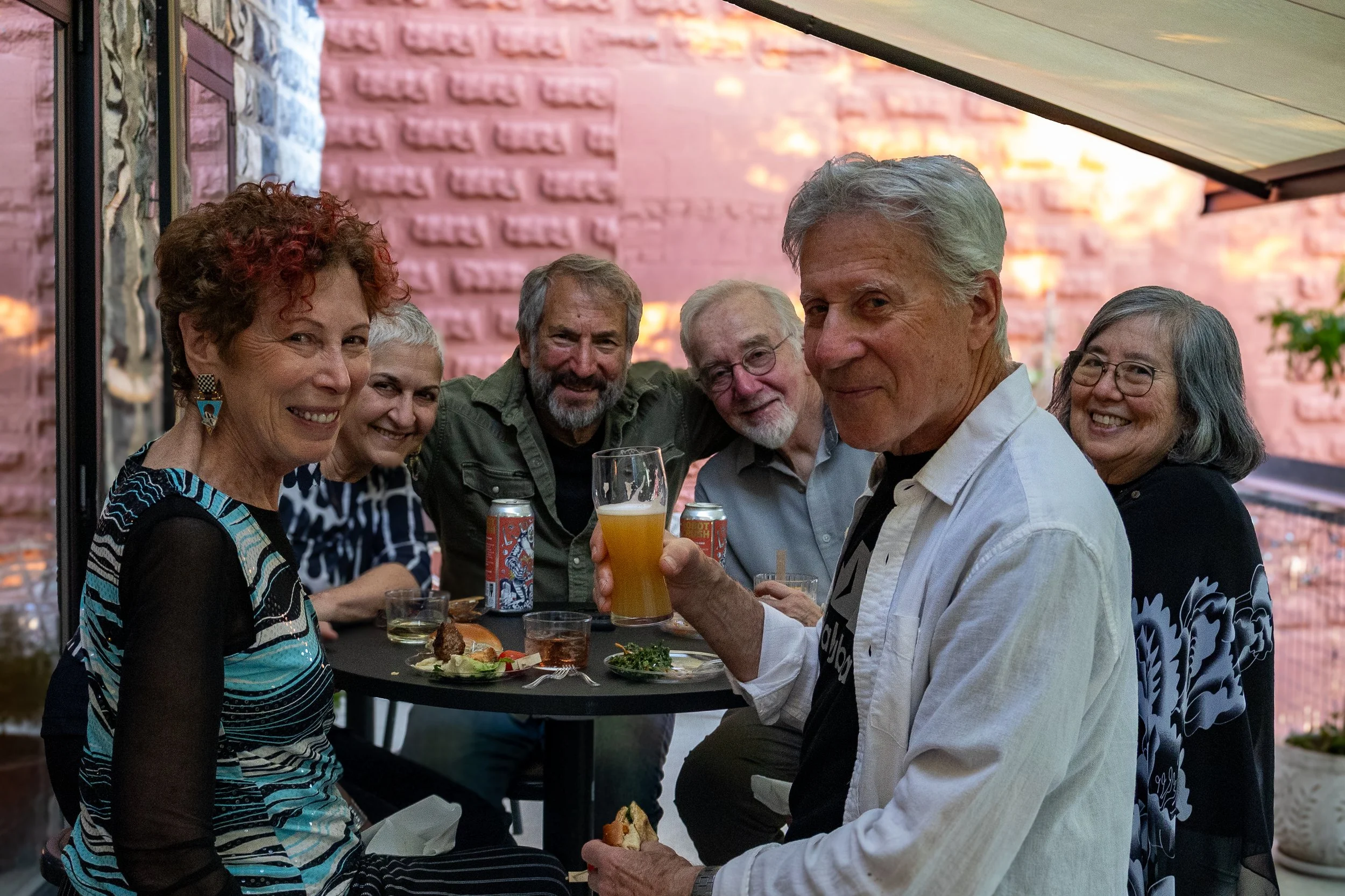 A group of six smiling adults enjoying a meal together at an outdoor gathering, with food and drinks on the table, and a pink brick wall in the background.