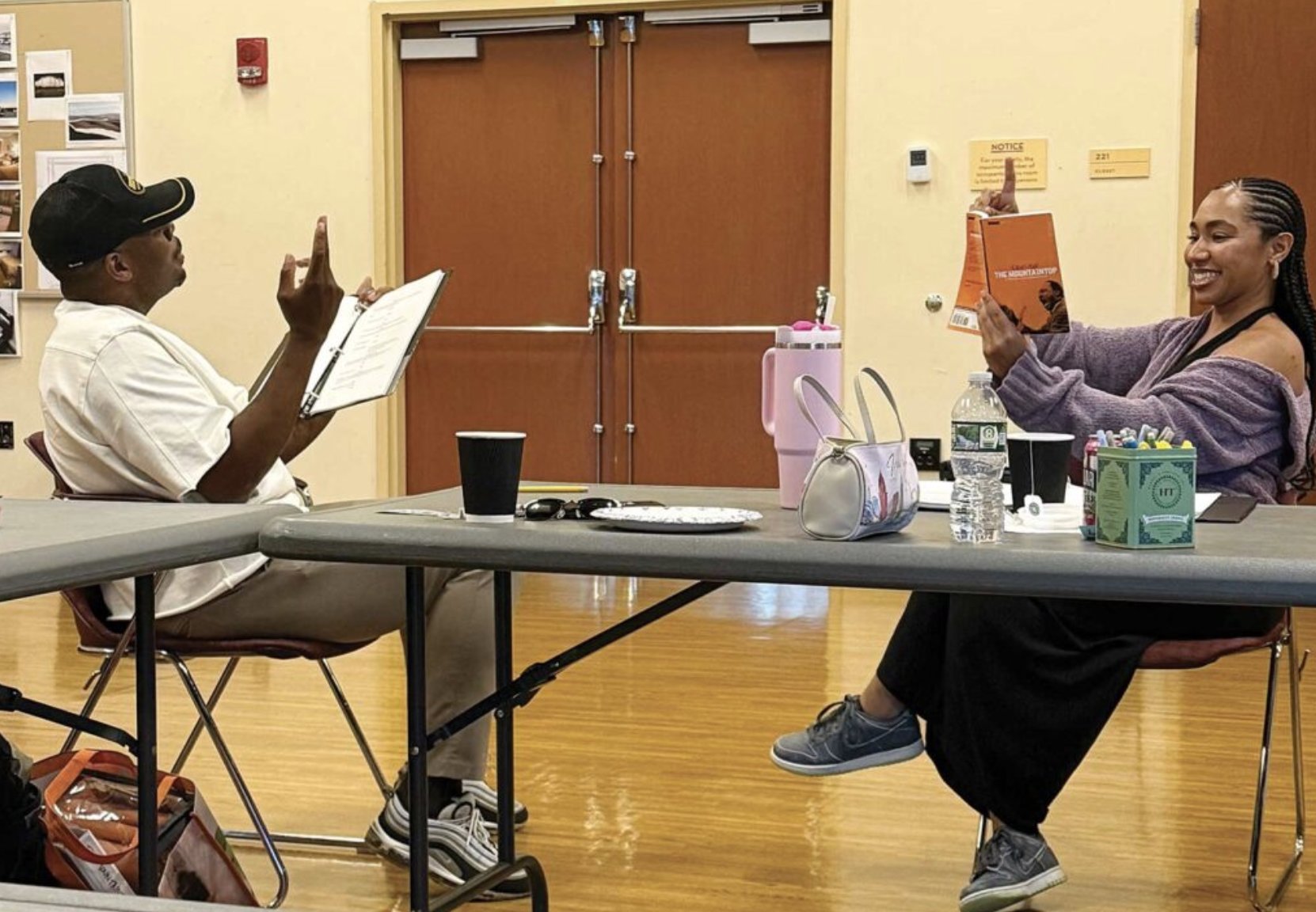 Two people seated at a table in a room with yellow walls, engaging in a conversation. The man on the left is wearing a white shirt and a black cap, holding a book and gesturing with his right hand. The woman on the right is wearing a purple cardigan and black pants, smiling, and holding a book. The table has various items including a water bottle, a purse, a black cup, and pens.