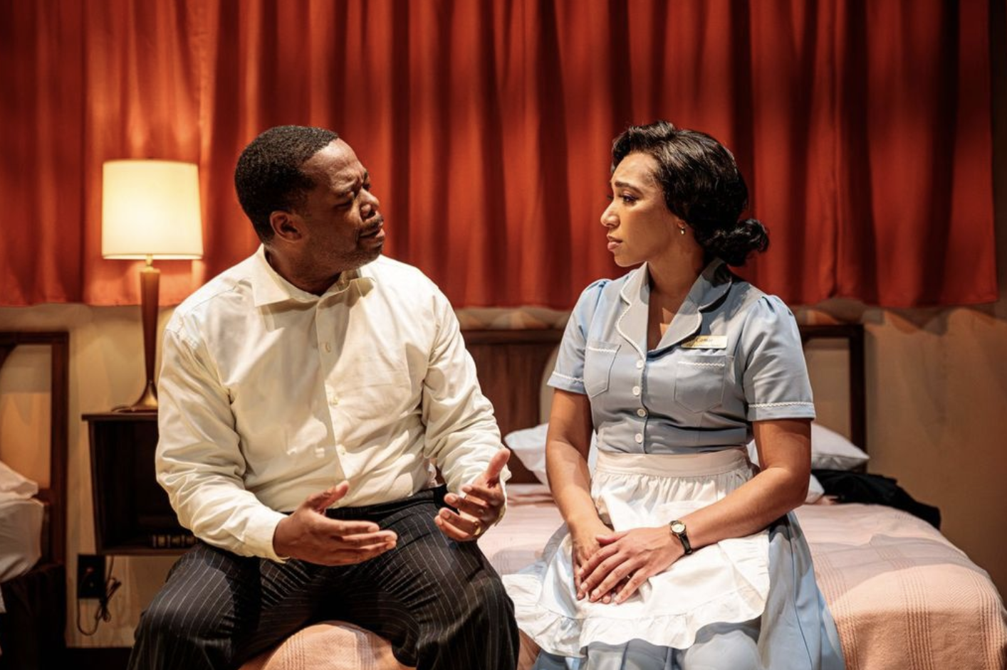 A man and woman sit on a bed having a serious conversation in a room with red curtains and a lamp.