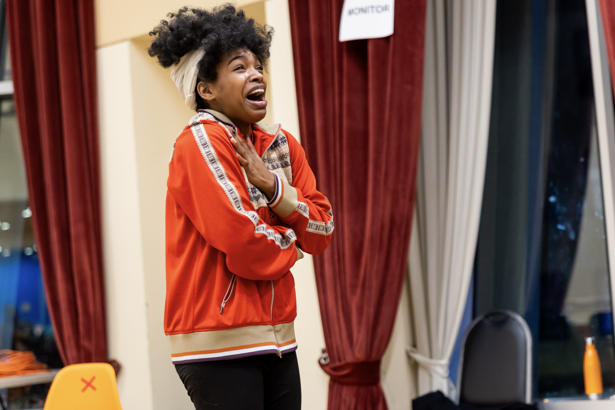 A woman with curly hair wearing a orange jacket is seen clutching her throat and appearing to cry or scream in a room with red curtains and colorfully decorated setting.