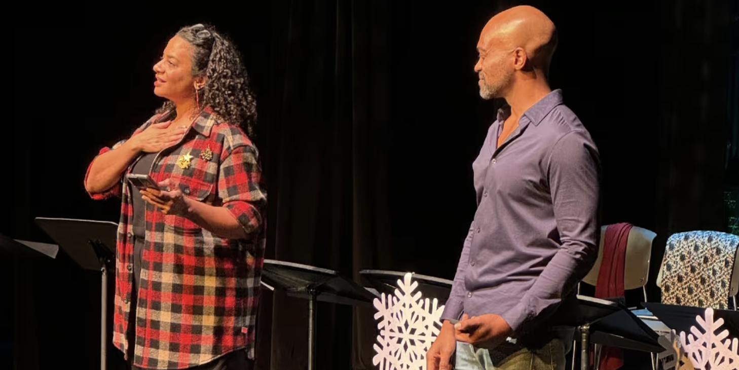 A woman with long, curly gray hair wearing a red and black plaid shirt, standing with her hand on her chest, holding a phone, and a man with a bald head and a gray beard, wearing a purple shirt, standing nearby with his hand in his pocket during a stage performance.