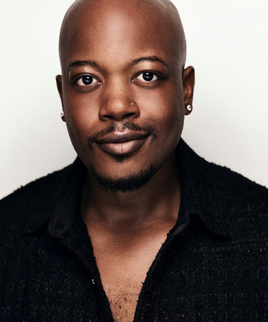 A close-up portrait of a bald Black man with earrings and a nose ring, wearing a black shirt, looking directly at the camera against a plain light background.