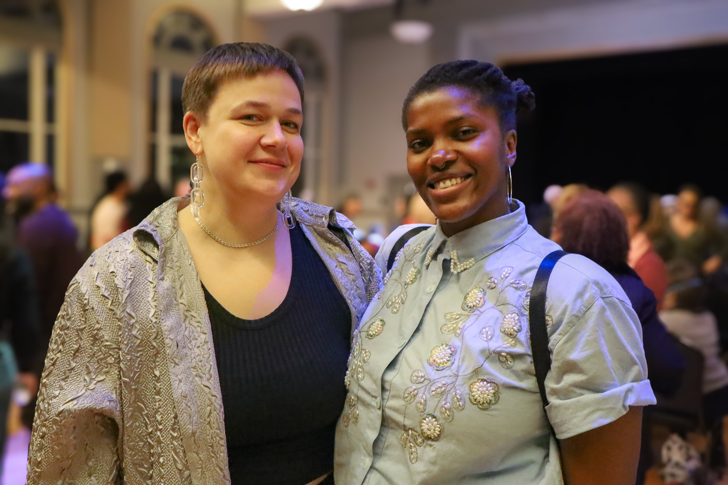 Two women standing together at an indoor event, smiling at the camera. One woman has short brown hair, fair skin, and is wearing a textured beige jacket and large earrings. The other woman has short black hair, dark skin, and is wearing a light-color