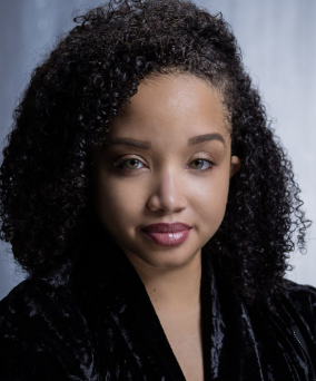 Close-up portrait of a young woman with curly dark hair, wearing a black top, looking at the camera with a neutral expression against a plain background.