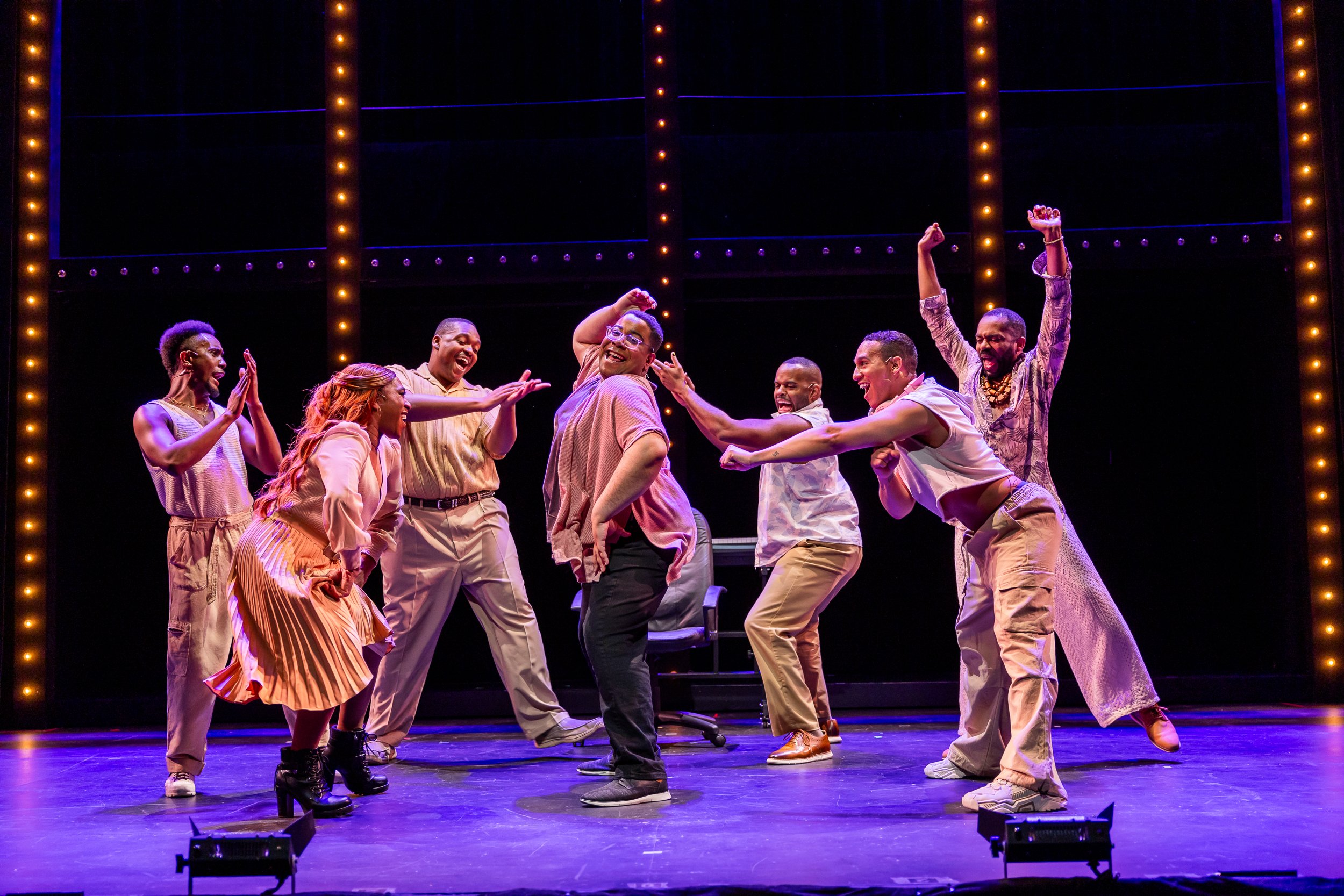 A group of people performing a lively dance on stage under bright lights. They are smiling and engaging with each other, wearing casual, colorful clothing.