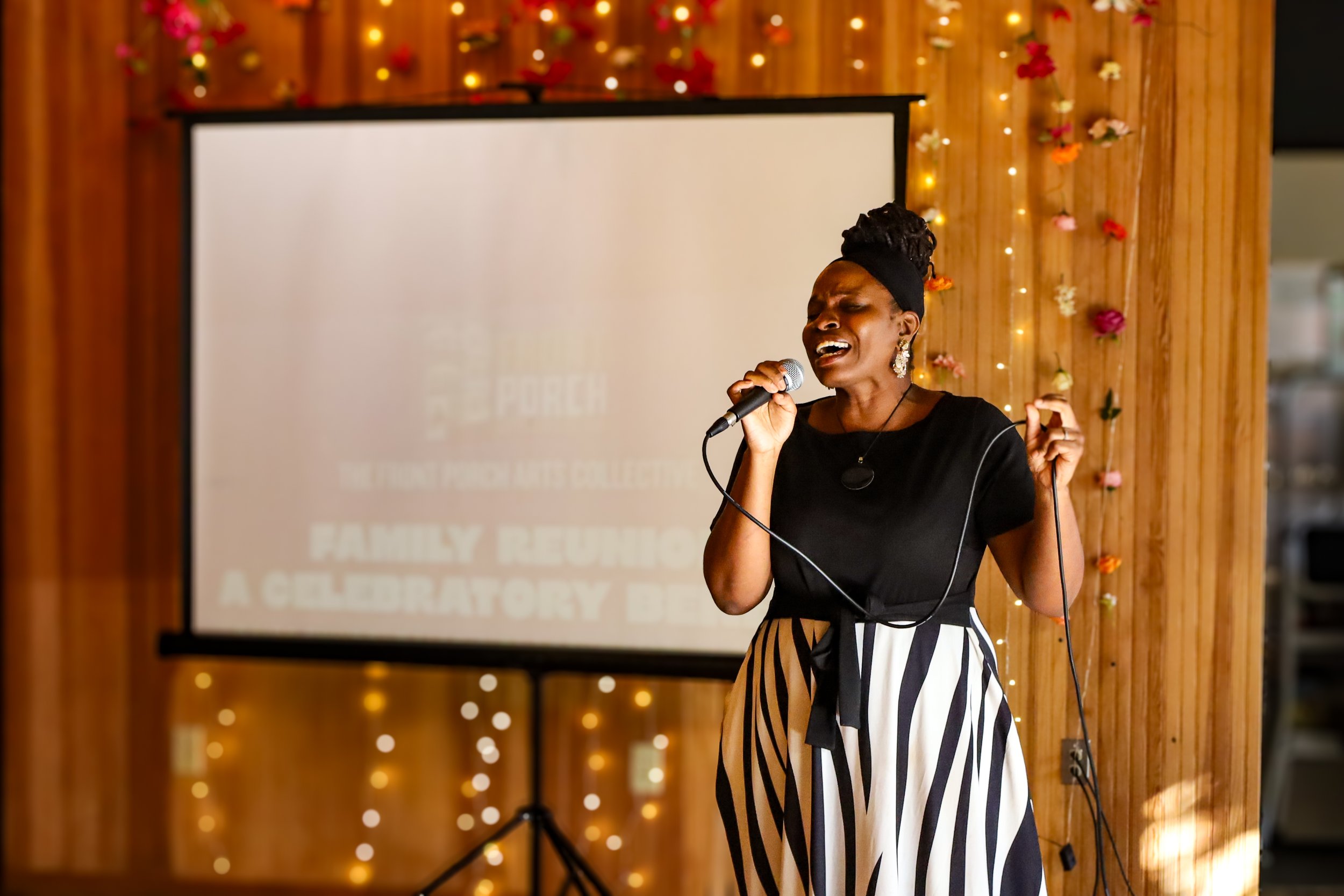A woman singing into a microphone during a performance at an indoor event with wooden walls and festive string lights and flower decorations.