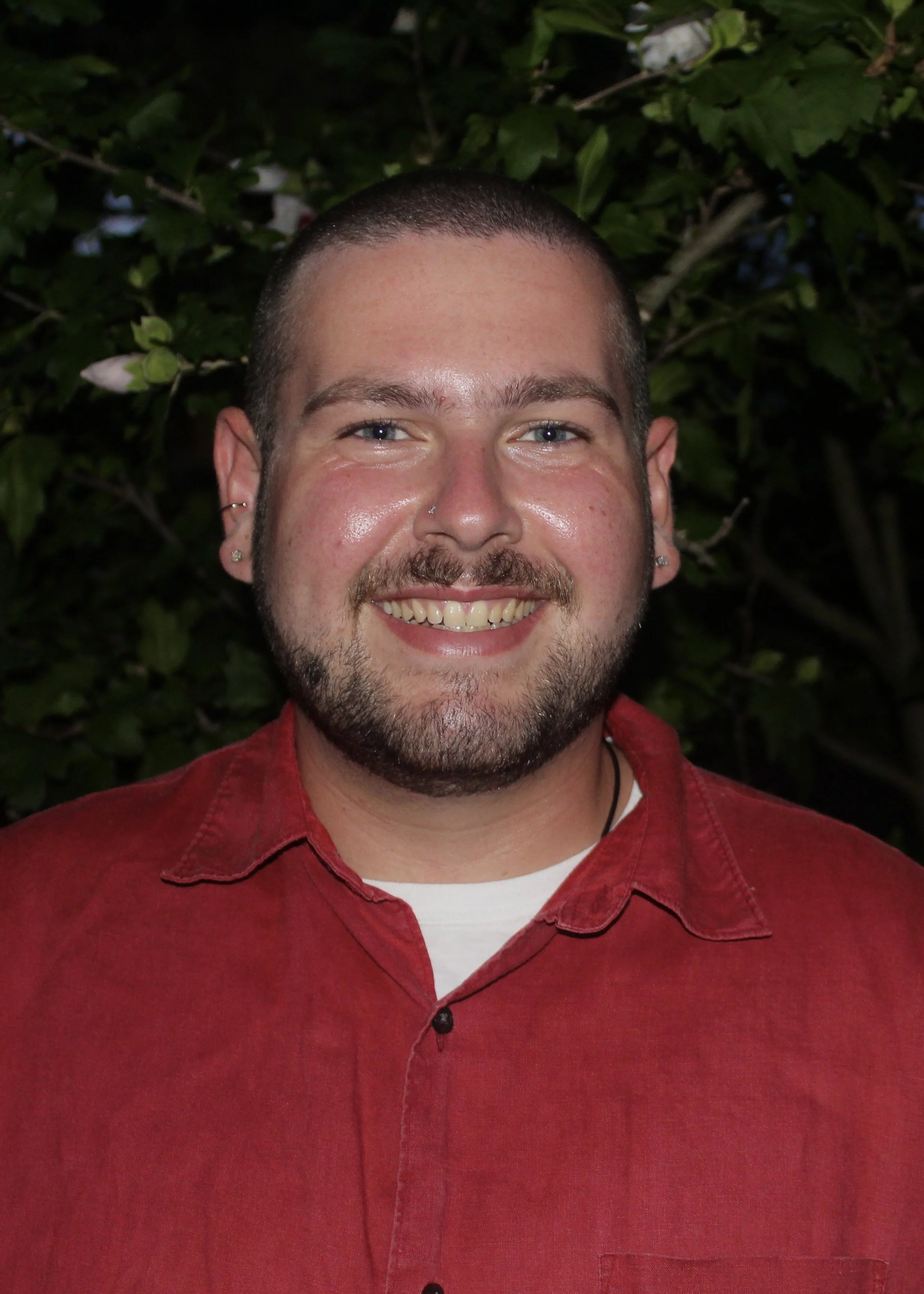 A smiling man with a beard and short hair, wearing a red button-up shirt and earrings, standing in front of green leafy background.