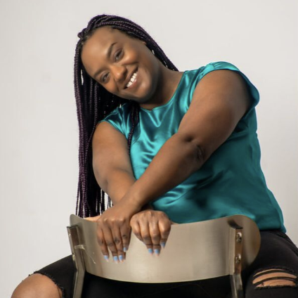 A woman with dark braided hair smiling while sitting on a chair, holding the backrest with both hands, wearing a shiny blue top and black ripped jeans.