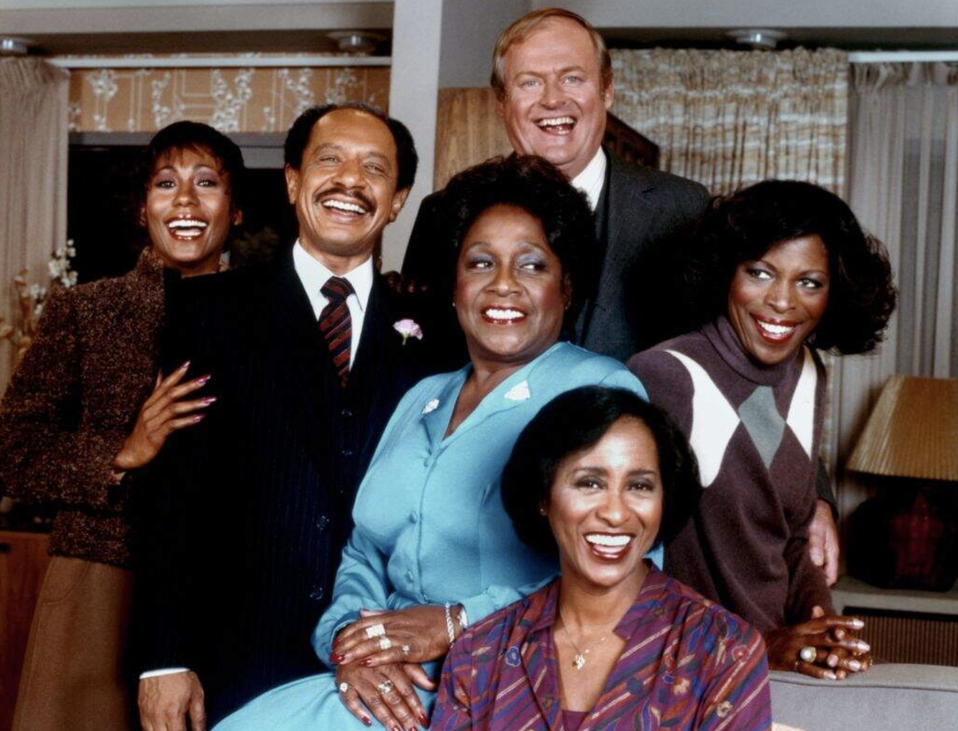 Six people smiling in a cozy indoor setting, posing together for a group photo, with a mix of men and women dressed in formal and casual attire.
