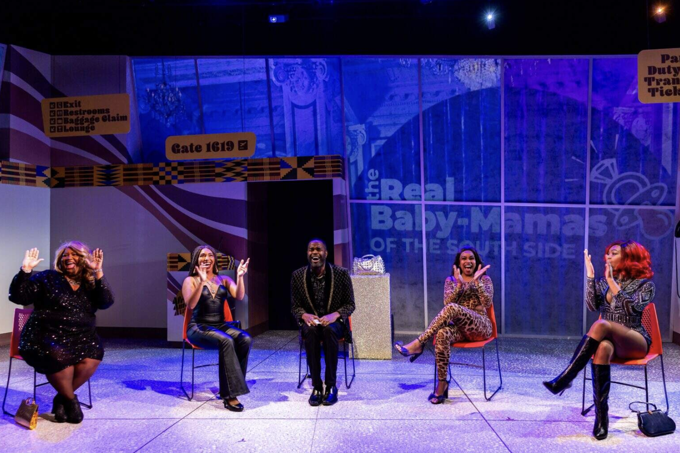 Five women are seated on chairs in a colorful, lively setting, laughing and enjoying themselves in a stage or studio environment with signage for the musical "The Real Baby.Mamas of the South Side."