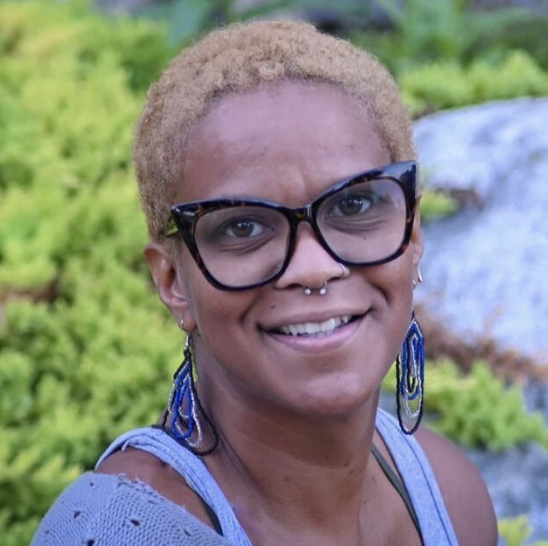 Close-up of a smiling woman with short, blonde hair, large black glasses, and blue earrings, outdoors with green foliage and rocks in the background.