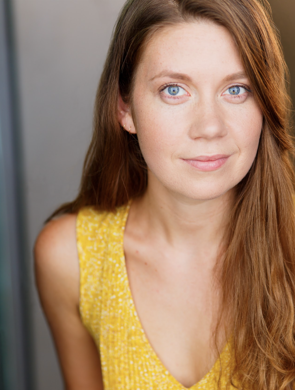 Close-up of a woman with long red hair, blue eyes, and light skin, wearing a yellow sleeveless top, looking at the camera with a subtle smile.