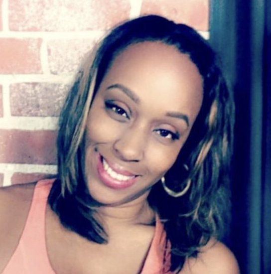 A smiling woman with shoulder-length hair, wearing hoop earrings and a pink tank top, standing against a brick wall.