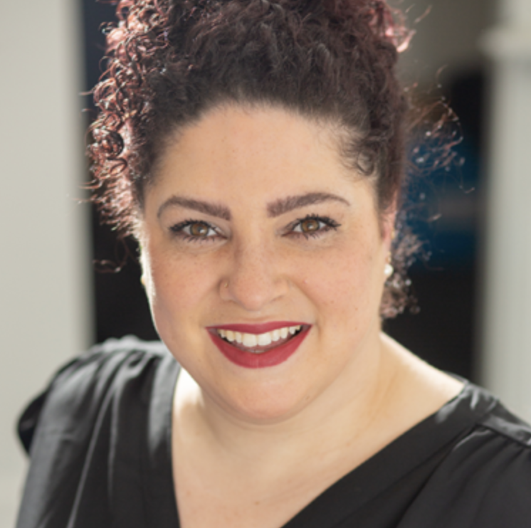 A woman with curly dark hair, light skin, green eyes, wearing red lipstick and pearl earrings, smiling at the camera.