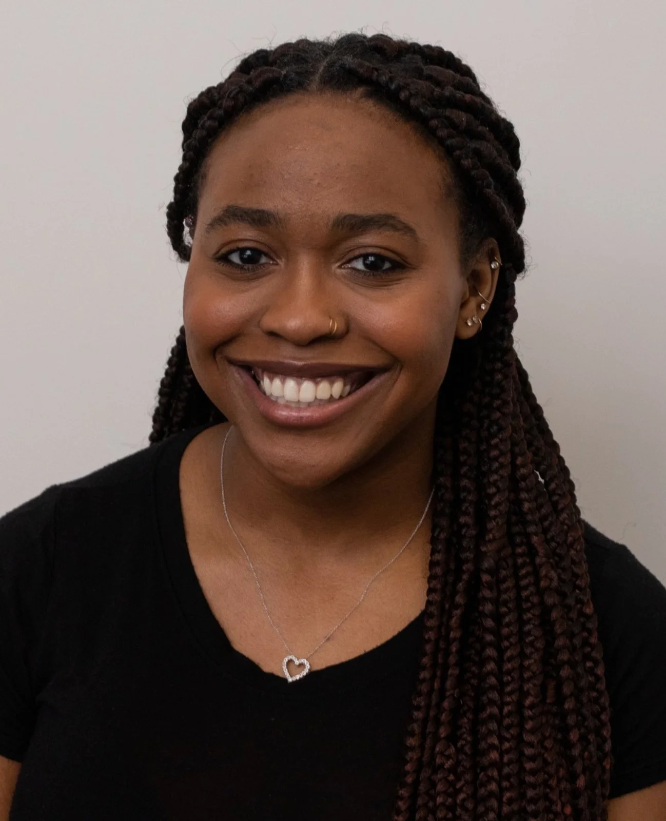 Close-up portrait of a smiling woman with braided hair, wearing a black shirt and a silver heart-shaped necklace, standing against a plain white background.