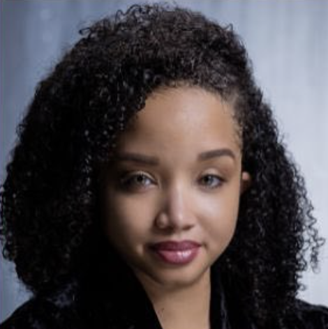Close-up of a young woman with curly black hair and light skin, smiling softly, looking at the camera.