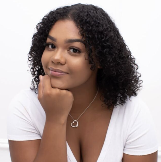 A woman with curly hair resting her chin on her hand, wearing a white top and a silver necklace with a heart pendant, smiling softly against a plain white background.