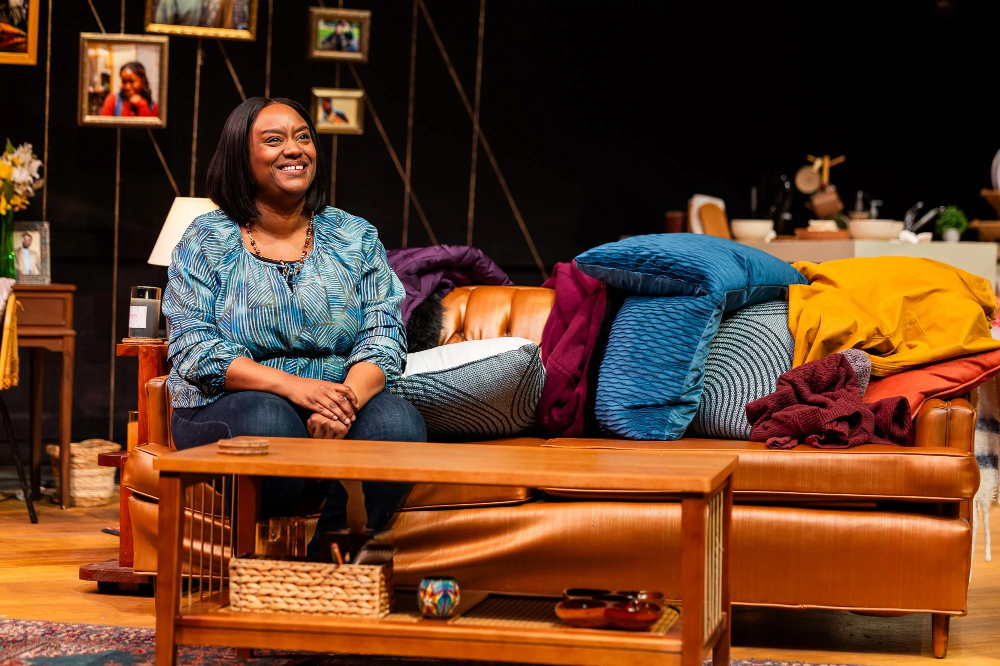 A woman with shoulder-length black hair, wearing a blue patterned blouse, sitting on a brown leather couch in a cozy living room. The background features picture frames on the dark wall and a cluttered side table. The coffee table in front has some small objects, and blankets or clothes are piled on the couch.