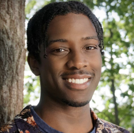 A smiling young man with dark braided hair posing outdoors near a tree, with green leafy background.