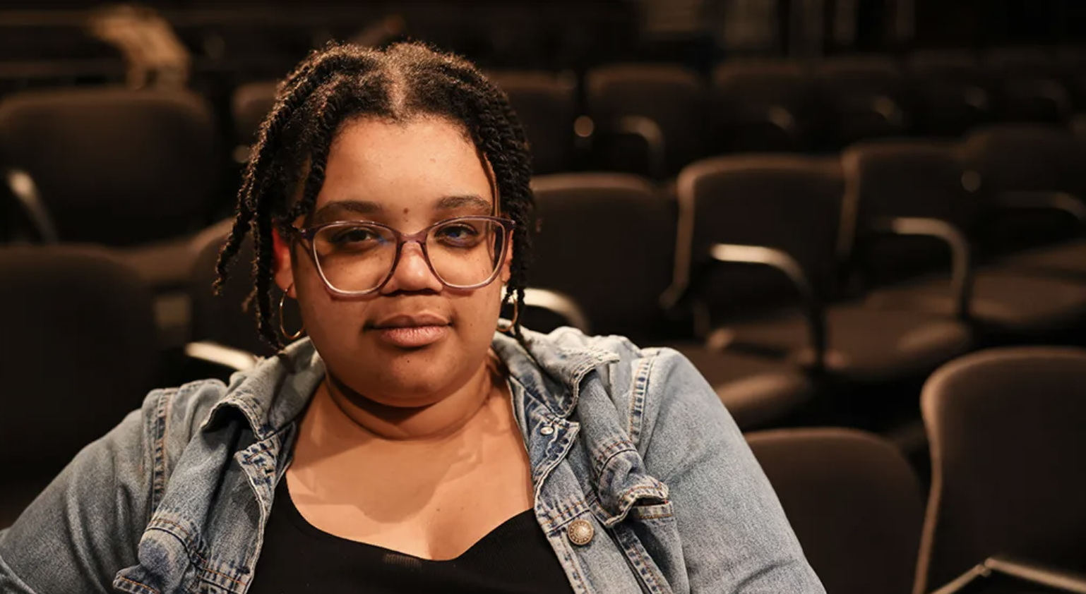 A woman with glasses and earrings sitting in an empty auditorium or theater with rows of black chairs.