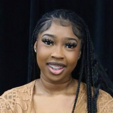 A young woman with braided hair, wearing gold earrings and a gold top, smiling at the camera against a dark background.