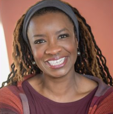 Portrait of a smiling woman with dreadlocks wearing a gray headband, pearl earrings, and a maroon and orange top against a blurred background.