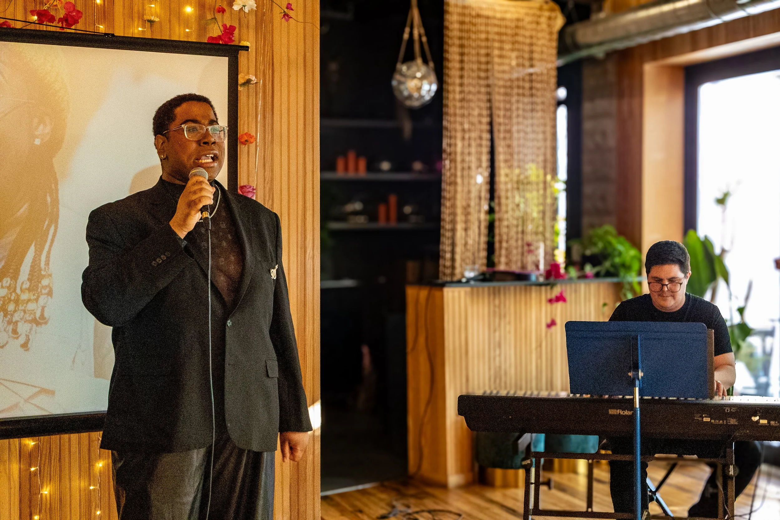 An individual in a black suit and glasses is holding a microphone and speaking in front of a projection screen in a warmly lit room. To the right, a person is seated at a keyboard, playing music, with large windows and decorative plants in the backgr