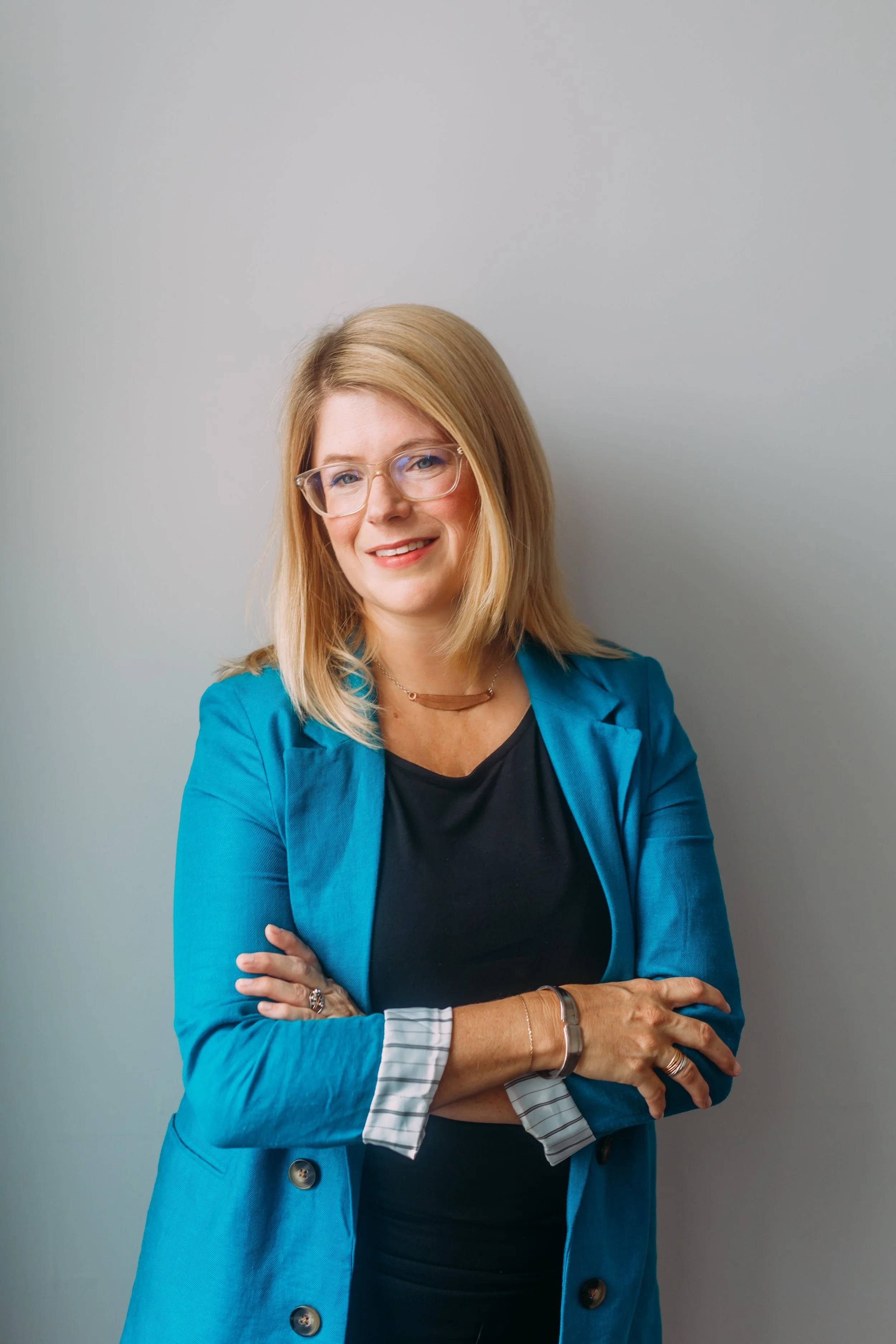 A woman with blonde hair, glasses, and a smile, is wearing a blue blazer over a black top, and has jewelry on her left hand and right wrist, standing against a plain light gray wall.