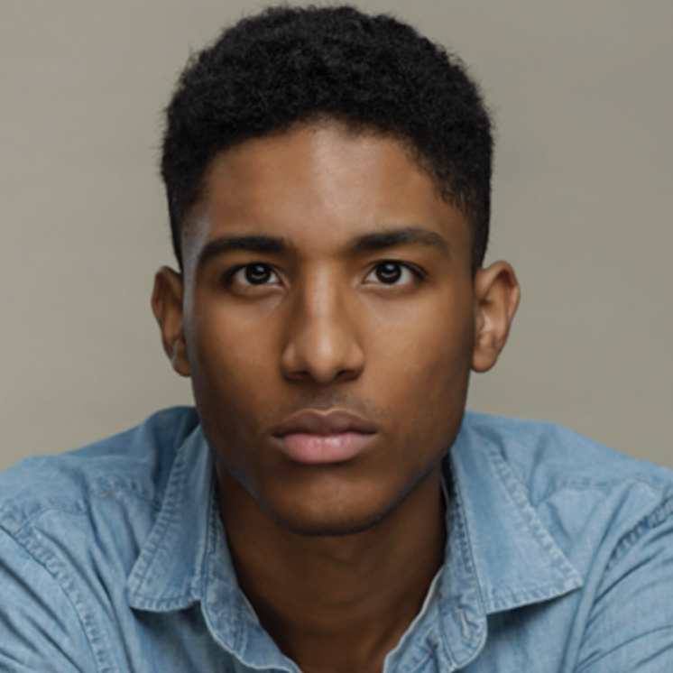 A young man with short curly black hair and a serious expression, wearing a light blue denim shirt, posed against a plain background.