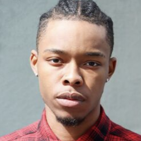 Close-up of a young man with braided hair and earrings, wearing a red plaid shirt, against a gray background.