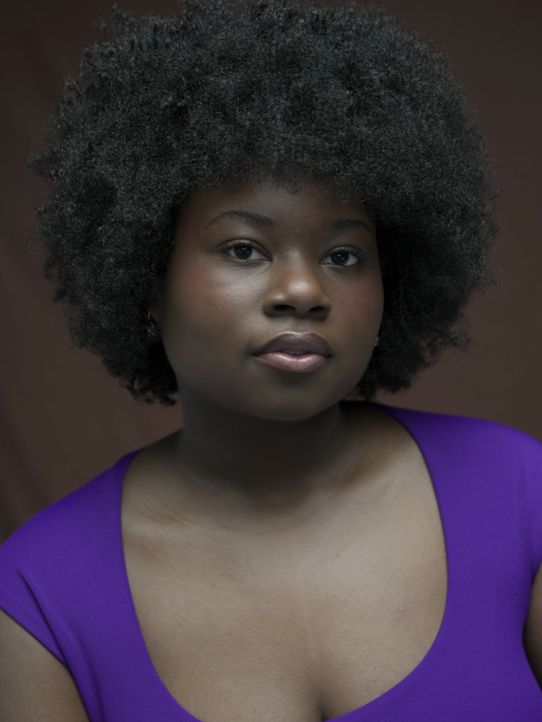 A woman with dark skin and a large afro hairstyle, wearing a purple top, looking directly at the camera with a neutral expression.
