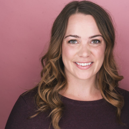 A woman with wavy brown hair and a bright smile, wearing a dark purple top against a pink background.