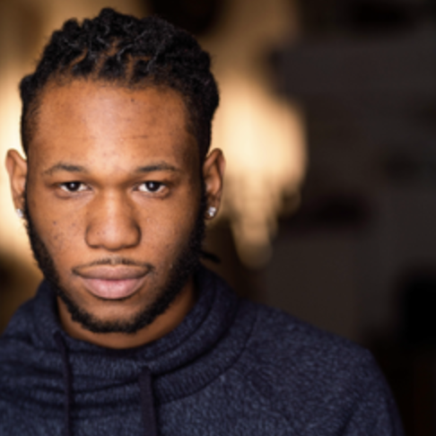 Close-up portrait of a young man with dark skin, short braids, piercing eyes, and a slight smile, wearing a dark hoodie.