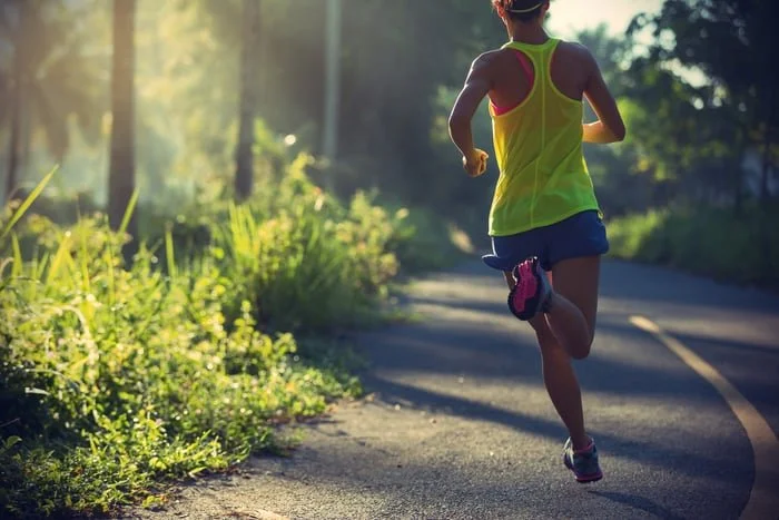 A woman running on a paved country road surrounded by greenery and trees in the morning sunlight.