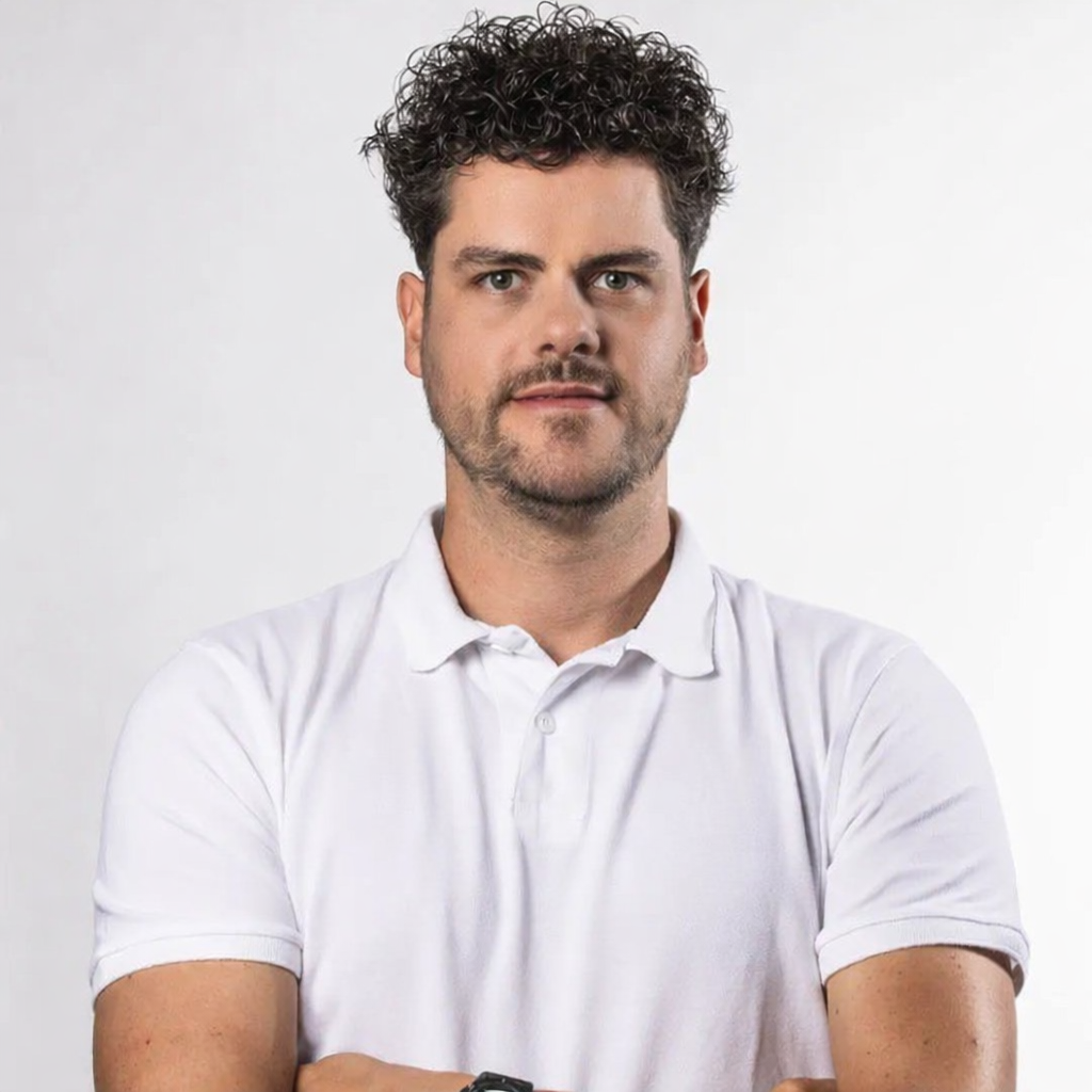 A man with curly dark hair and a beard wearing a white polo shirt, standing against a plain light background.