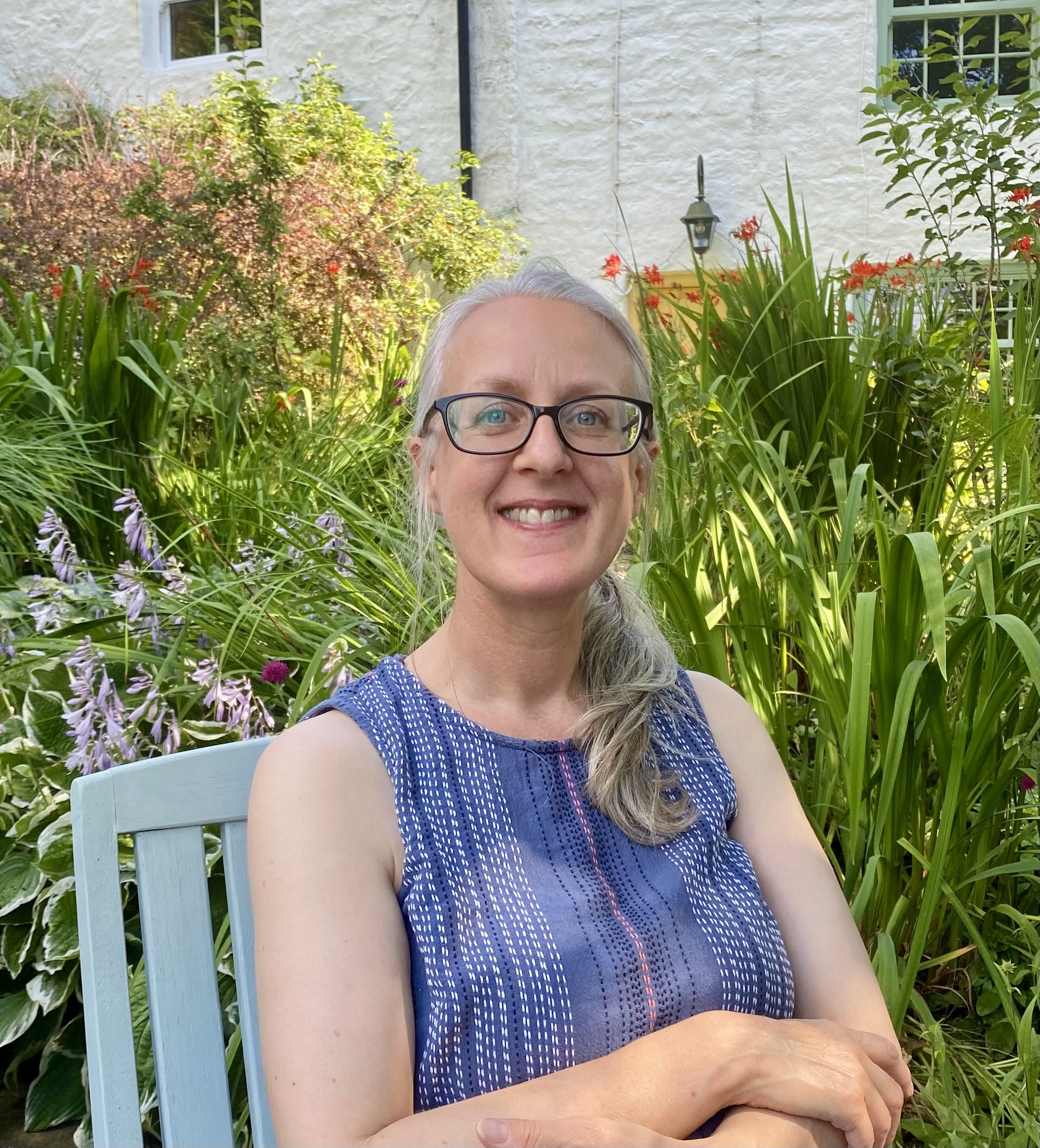 A smiling woman with gray hair and glasses sitting on a light blue chair in a garden with lush green plants and colorful flowers.