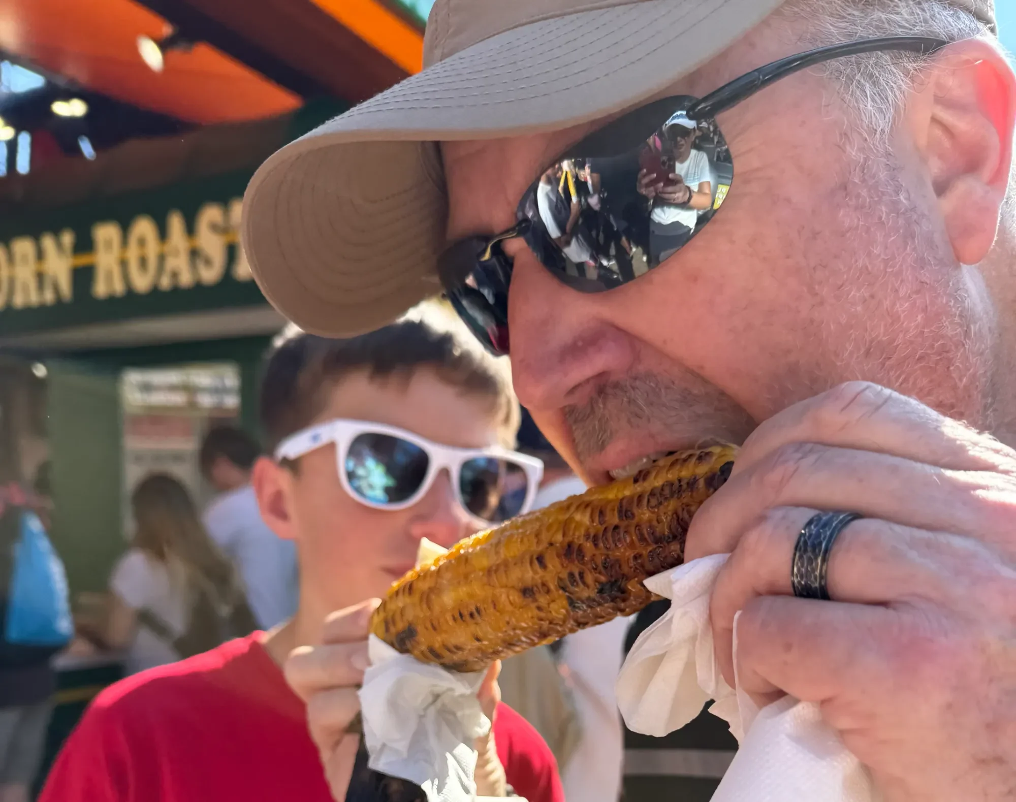 A man wearing a beige hat and sunglasses is eating roasted corn on the street. There are people in the background, and a green sign with yellow text is partially visible.