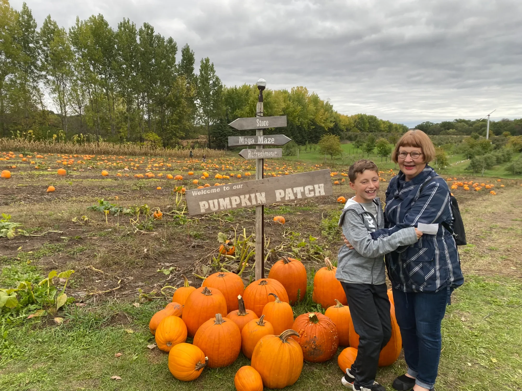 A woman and a boy smiling and hugging in front of a pumpkin patch with pumpkins, a sign that reads "Welcome to our Pumpkin Patch," and a backdrop of fields, trees, and cloudy sky.