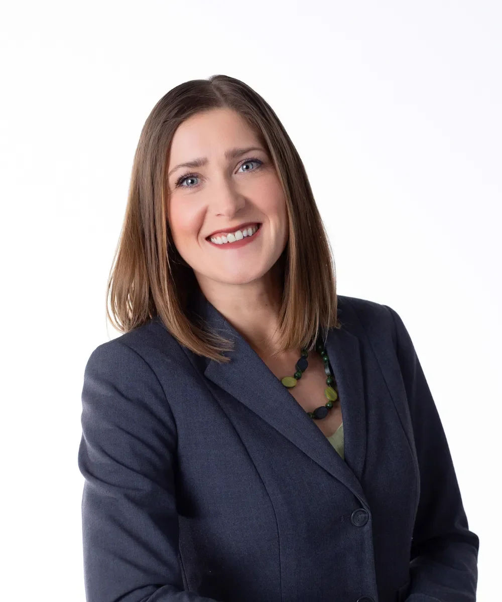 A woman with shoulder-length brown hair, blue eyes, and fair complexion, smiling, wearing a dark blazer and a multi-colored necklace, against a plain white background.