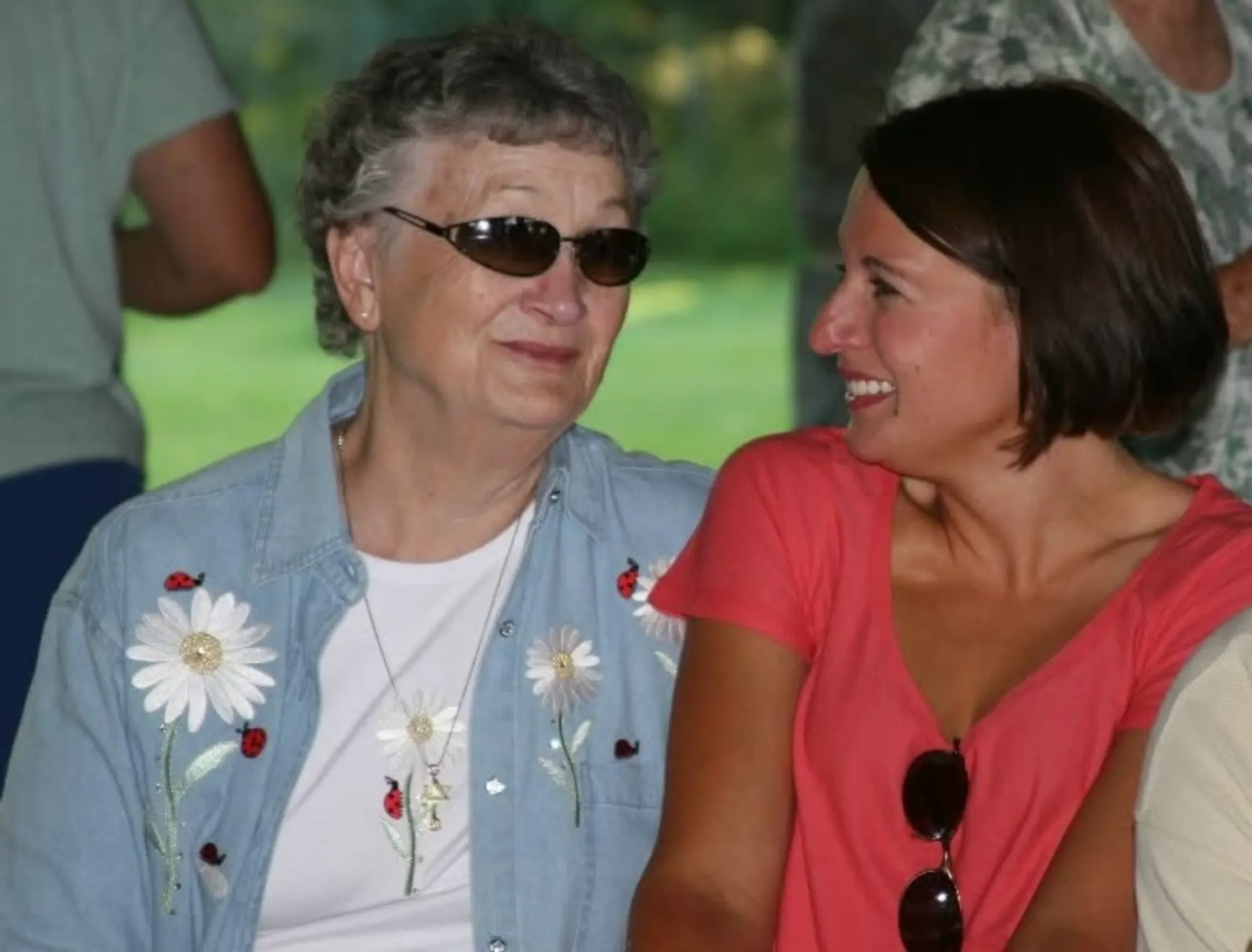 Senior woman with short gray hair and sunglasses smiling at a young woman with dark hair and a bright red shirt, in an outdoor setting with a blurred background.