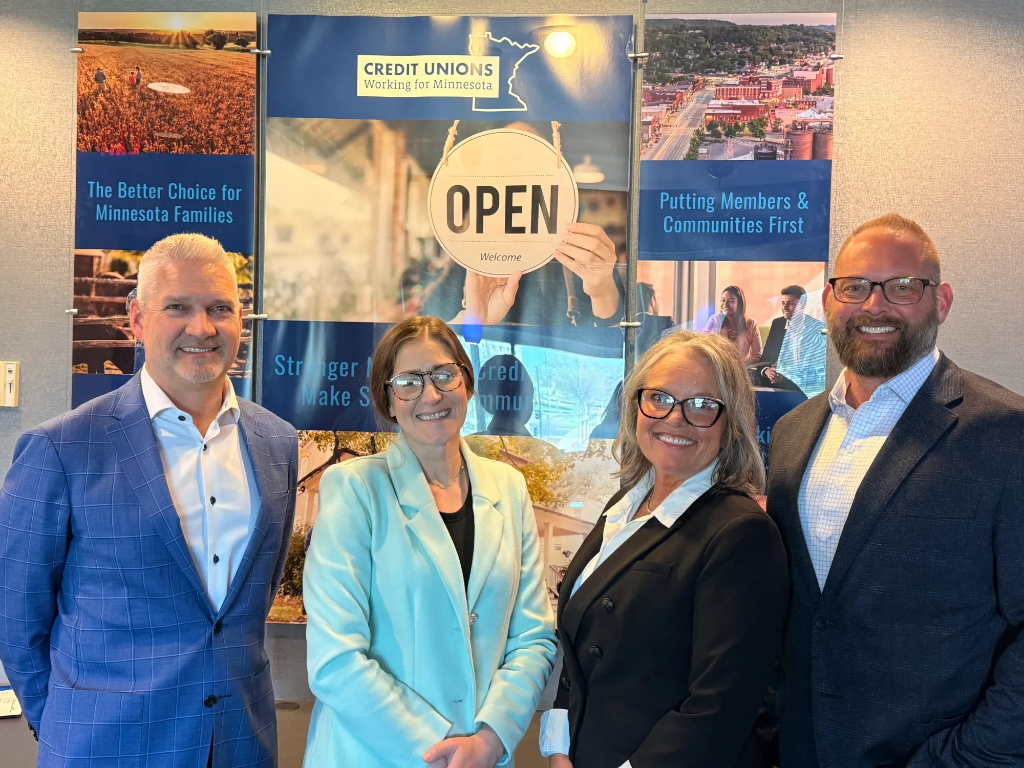 Group of four people standing together in front of a wall with Minnesota-themed posters and signs for a credit union event. The posters include images of Minnesota landscapes, business districts, and community scenes.