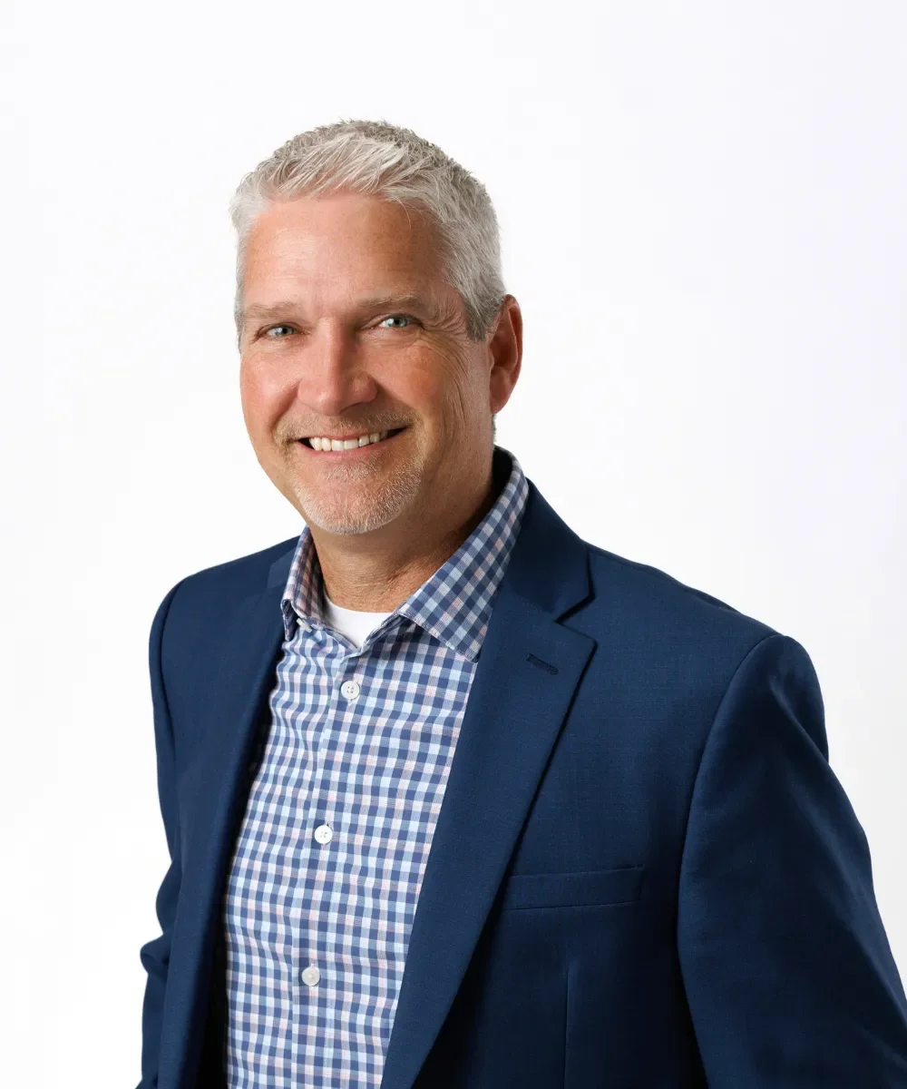 A middle-aged man with short gray hair and a beard, smiling, wearing a dark blue blazer over a checkered blue and white shirt, against a plain white background.