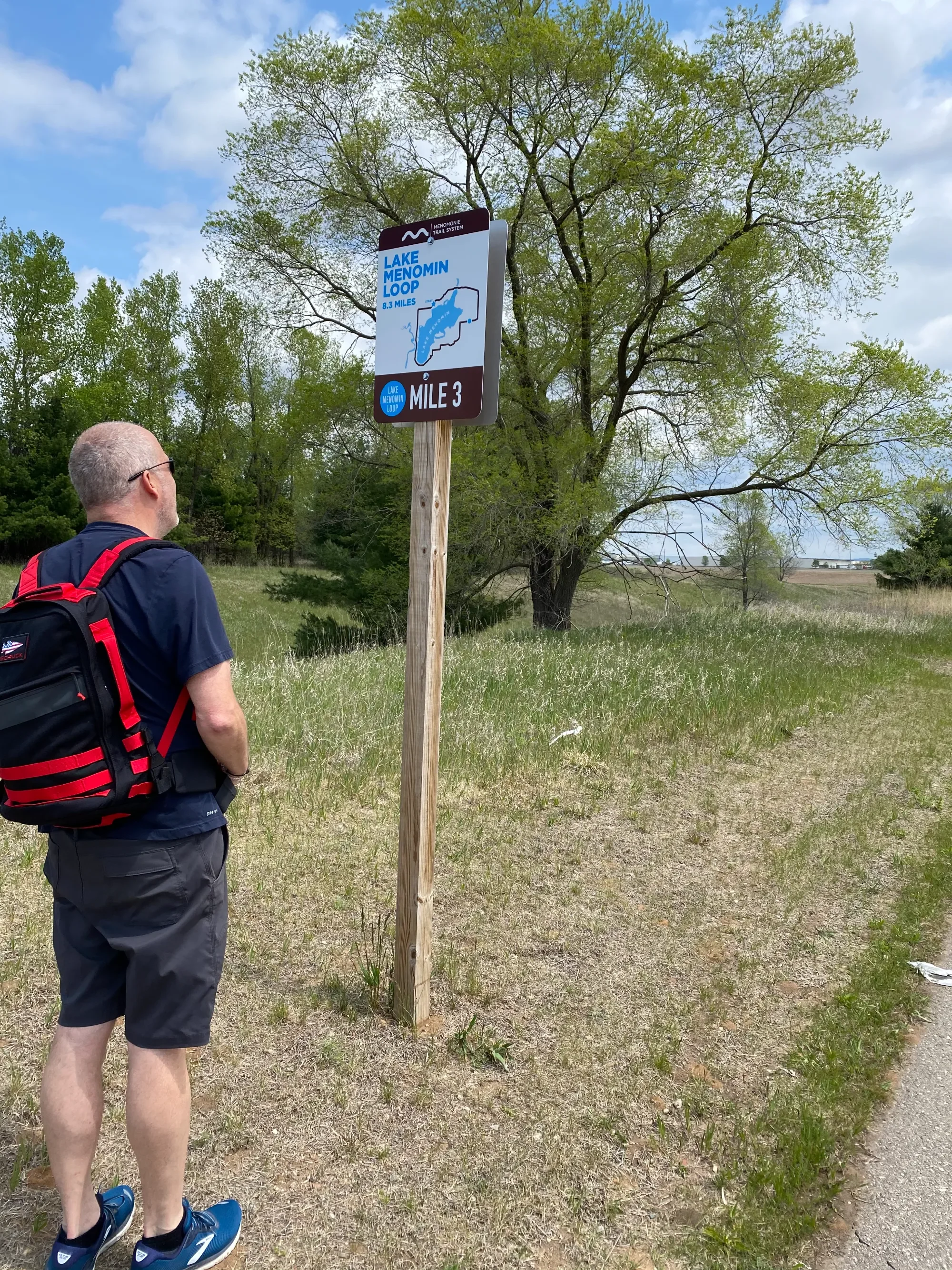 A man with a gray beard and sunglasses wearing a navy shirt, black shorts, blue sneakers, and a red and black backpack stands outdoors in front of a sign for Lake Menomin Loop, which indicates the trail is 8.3 miles long and the man is at the 3-mile mark. The background features green trees and a partly cloudy sky.