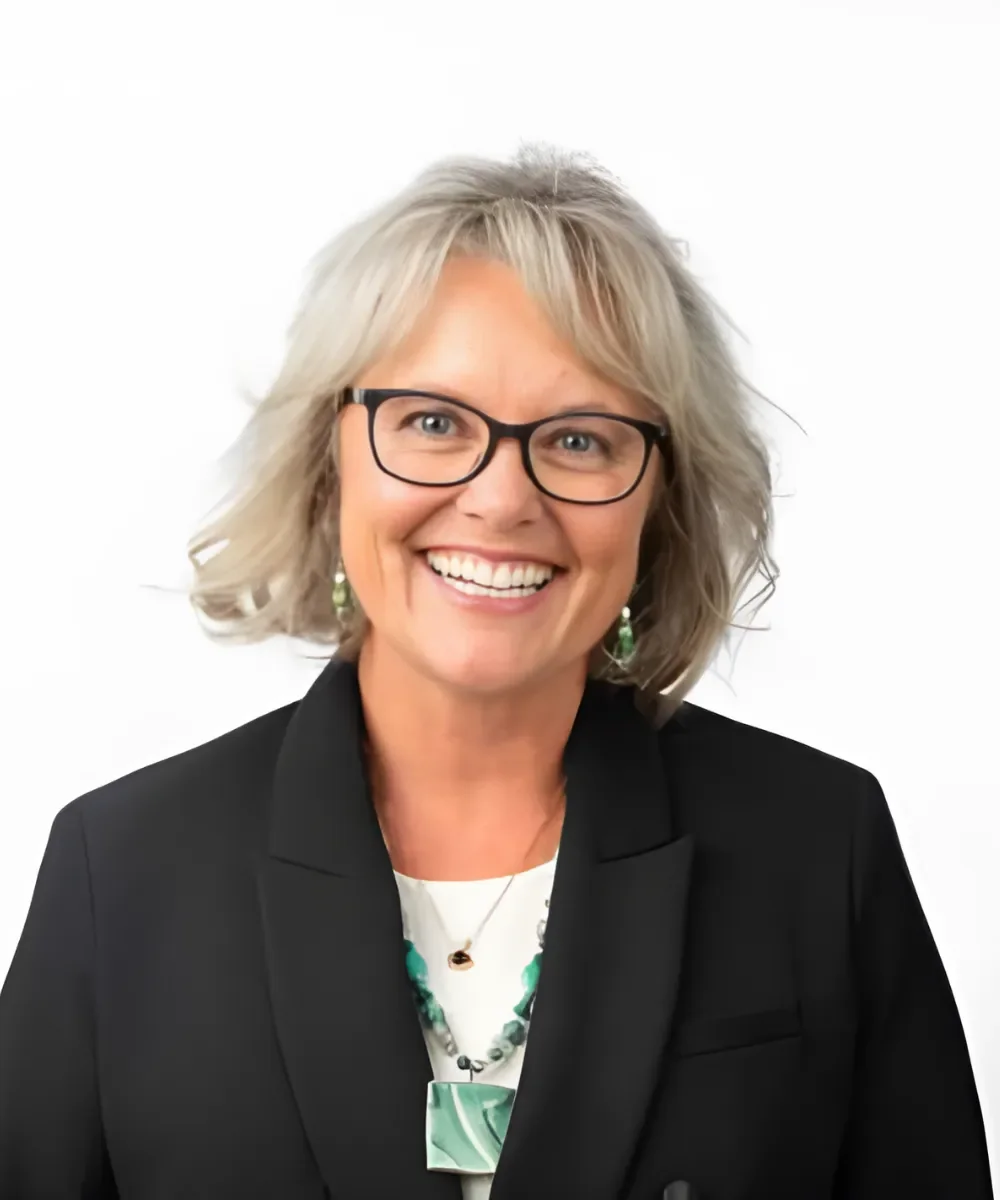 A smiling woman with short gray hair, black glasses, wearing a black blazer, patterned necklace, and earrings, against a white background.
