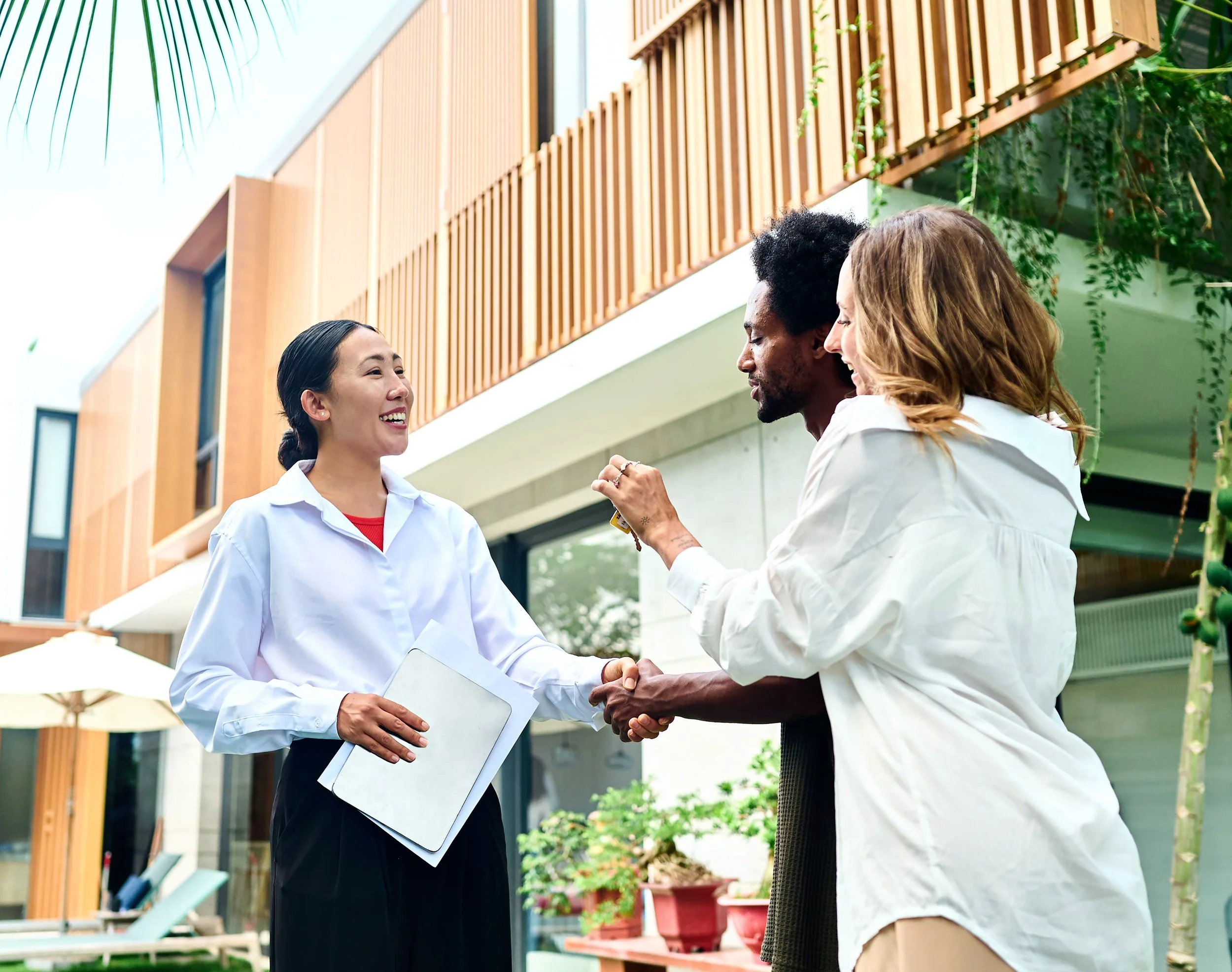 Three people exchanging a handshake outside a modern building, smiling and holding documents. One woman has black hair tied back, another woman with curly red hair, and a man with afro hairstyle.