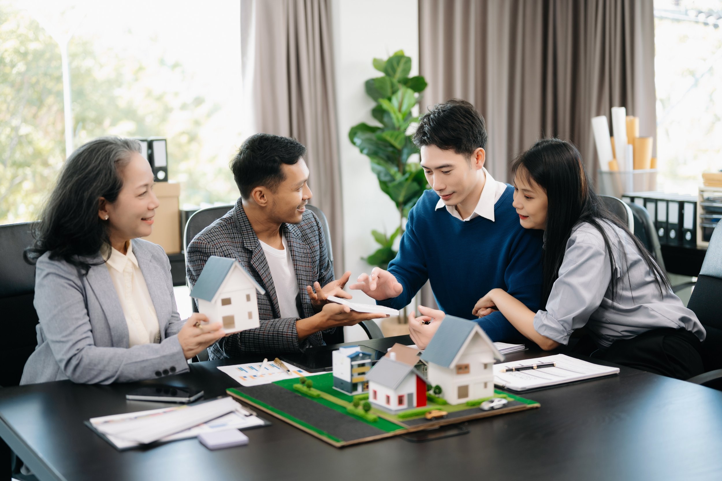 Meeting of five people discussing real estate models and plans, sitting around a table with miniature houses and documents.